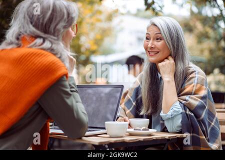 Mature Asian lady and grey haired friend with laptop sit together on outdoors cafe terrace cafe Stock Photo