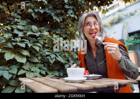 Cheerful senior woman with spectacles eats strawberry dessert sitting at table on outdoors cafe terrace Stock Photo