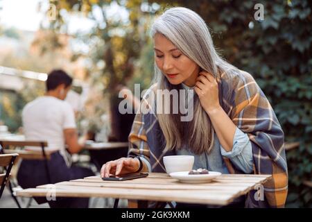 Long haired mature Asian lady reads message on mobile phone sitting on outdoors cafe terrace Stock Photo