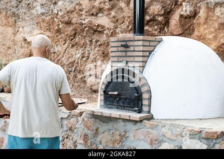 Man putting pizza in handmade white painted wood oven built outdoors with shovel Stock Photo