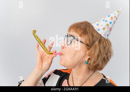 Portrait of a smiling elderly woman in a festive cap holding a whistle tongue on a white background Stock Photo