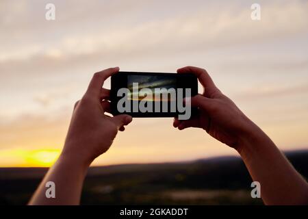Closeup of hands of young female traveller holding smartphone taking picture of beautiful morning sunrise Stock Photo