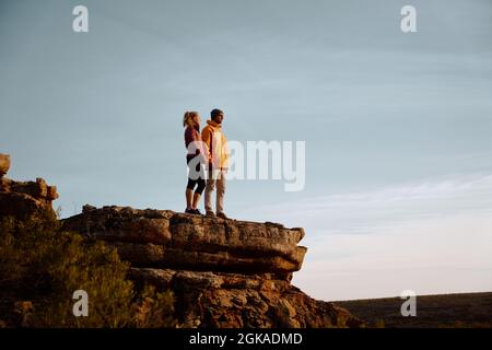 Young couple holding hands looking away standing at peak of cliff after successful hike Stock Photo