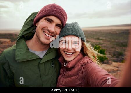 Portrait of cheerful young woman embracing man in winter clothing taking selfie during camping on mountain Stock Photo