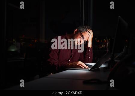 Tired and worried businessman at workplace in office holding his head on hands after late night work on laptop Stock Photo