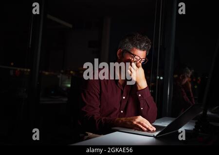 Tired middle age businessman rubbing his eyes while sitting in front of laptop computer working on project till late night Stock Photo