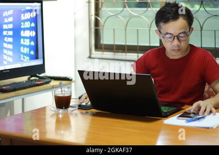 Asian man working from home.  Table in the room and window is behind. On the table have laptop, smartphone, coffee, paper and pen. There is big screen Stock Photo