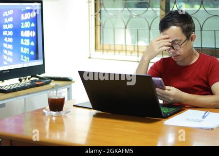 Asian man working from home, his hand hold the head that ben down because of stress with work . On the table have laptop, smartphone, coffee and paper. Stock Photo