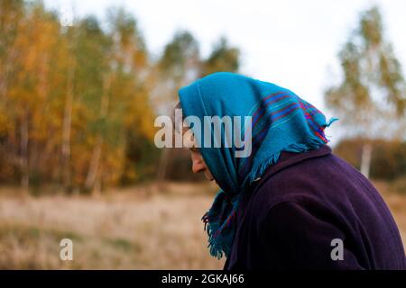 Defocus portrait of russian grandmother senior old woman standing in yellow autumn park. Old women in coat and shawl. Sad person. Nature background. O Stock Photo