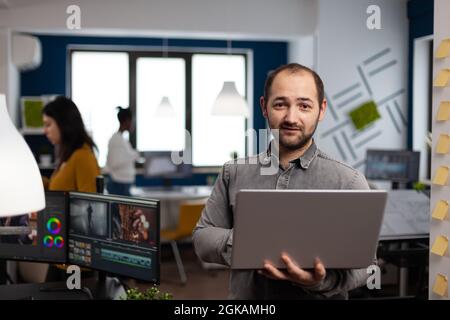creative video editor worker standing in front of camera smiling working in digital agency office holding laptop. Man videographer works in multimedia post production studio editing video content Stock Photo
