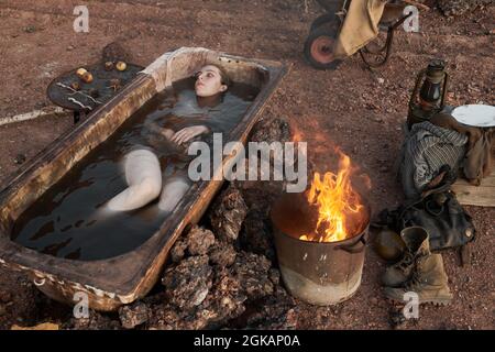 High angle view of depressed woman lying in the bath with dirty water outdoors and warming up near the fire Stock Photo