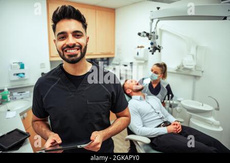 Caucasian male dentist holding digital tablet smiling with over watching female colleague working on male client  Stock Photo