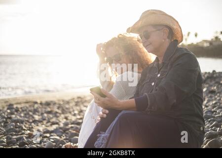 Senior woman with her young daughter relaxing on beach. Mother showing mobile phone to daughter and smiling. Old woman sharing media content with her Stock Photo