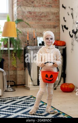 Little mummy holding pumpkin bucket for halloween treats while standing in domestic room Stock Photo