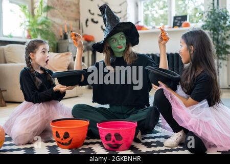 Young female in witch attire putting treats in hats of two adorable girls while sitting on the floor Stock Photo