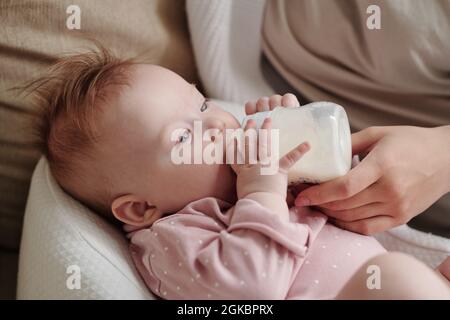 Cute toddler girl eating milk while holding bottle, young mother helping her Stock Photo