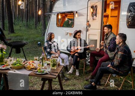 Friendly company sitting by house on wheels and singing songs while female in hat playing guitar Stock Photo