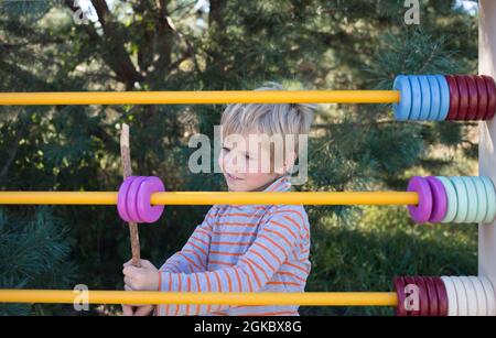 Little boy with wooden colorful abacus does math exercises outdoors, learning addition and counting. The preschooler child is studying. Back to school Stock Photo
