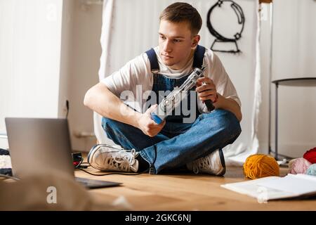 Young white man using laptop and sewing machine while working on craft rug at home Stock Photo