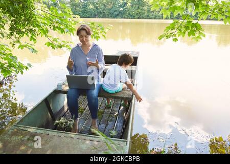 Single mother using laptop computer at lakeside in summer with son with her Stock Photo