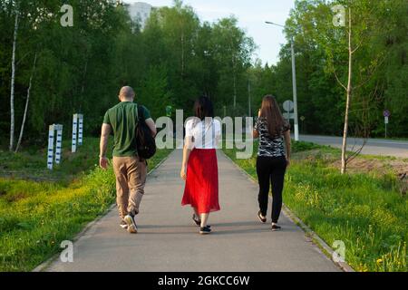 A guy and two girls are walking along the road. Young people are walking down the street. Three people shot from the back. Walk of young people around Stock Photo