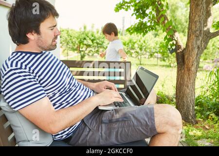 Man working on laptop computer in garden in summer with child in background Stock Photo