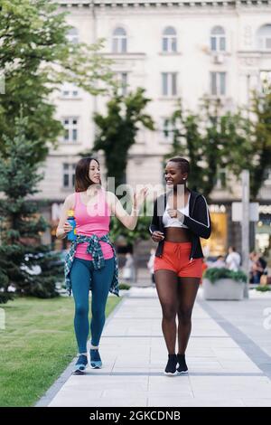Cheerful smiling friends in sportswear walking after a sport session in the city discussing. Multiethnic women having a fitness workout jogging. Stock Photo