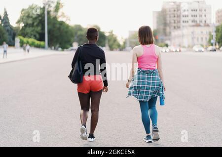 Friends back view in sportswear walking after a sport session in the city discussing . Multiethnic women having a fitness workout jogging. Stock Photo