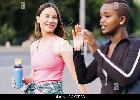 Cheerful smiling friends in sportswear walking after a sport session in the city discussing and having fun. Multiethnic women having a fitness workout jogging. Stock Photo