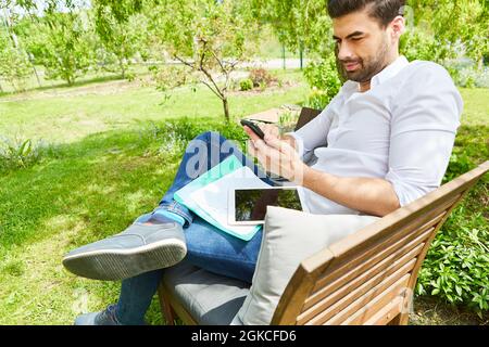 Young man as a freelancer writes a text message on the smartphone in the green garden Stock Photo