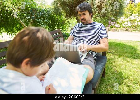 Single father on laptop computer in garden in summer with son Stock Photo