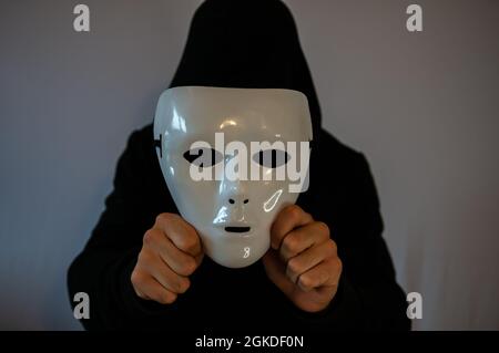 Man with black hood holding an anonymous white mask on dark background Stock Photo
