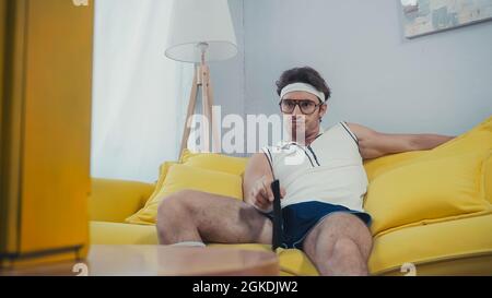 young bored man in eyeglasses and sportswear sitting on couch and switching channels Stock Photo