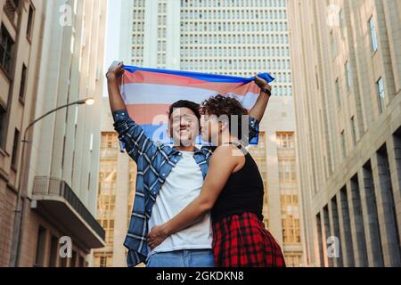 Gender nonconforming couple celebrating gay pride outdoors. Happy young queer couple raising the transgender flag while standing in the city. Young LG Stock Photo
