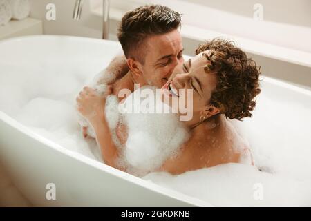 Happy young queer couple having fun in the bathtub. Romantic young couple enjoying a bubble bath together at home. Young LGBTQ+ couple spending qualit