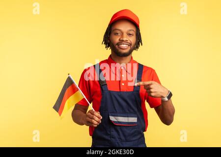 Satisfied handsome handyman in uniform with dreadlocks pointing finger at German flag in his hand and looking at camera with toothy smile. Indoor stud Stock Photo