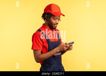 Side view portrait of positive deliveryman wearing uniform, using smartphone for taking new order for delivering, delivery service. Indoor studio shot Stock Photo