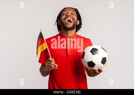 Screaming man wearing red casual T-shirt, football fan sincerely rejoicing, supporting favorite team holding ball and german flag, watching football. Stock Photo