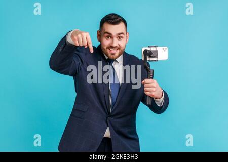 Businessman wearing official style suit, pointing down while communicating by video call or streaming, using steadicam, gesturing to subscribe. Indoor Stock Photo