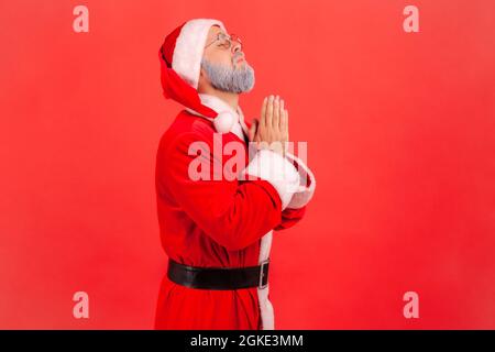 My God please help me. Side view of elderly man with gray beard wearing santa claus costume standing looking up and praying with worry face. Indoor st Stock Photo