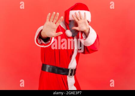Elderly man with gray beard wearing santa claus costume closing eyes with hand and stretching hand, moving in darkness eyesight problems. Indoor studi Stock Photo