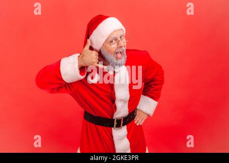 Excited elderly man with gray beard wearing santa claus costume making telephone gesture near ear and looking at camera with open mouth. Indoor studio Stock Photo