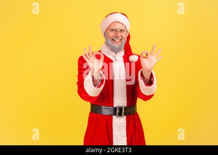 Portrait of satisfied elderly man with gray beard wearing santa claus costume showing okay sign with both hands, looking at camera toothy smile. Indoo Stock Photo
