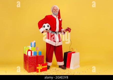 Full length of elderly man with gray beard wearing santa claus costume has satisfied expression, holding joystick and soccer ball, standing near gifts Stock Photo