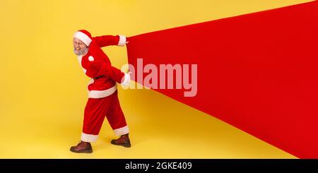 Full length portrait of elderly man with gray beard in santa claus costume showing big copy space for advertisement, carrying large bag with presents. Stock Photo