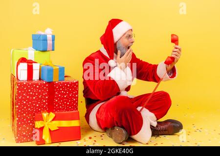 Surprised elderly man wearing santa claus costume sitting on floor with retro land line phone, Christmas congratulations, covering mouth with hand. In Stock Photo