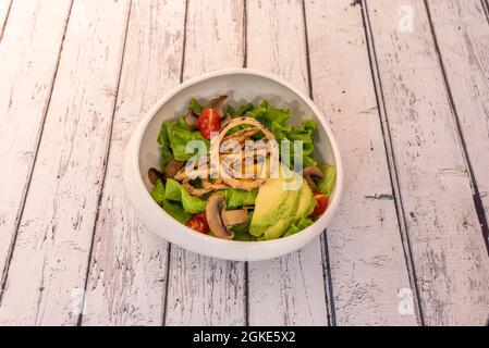 Avocado and mango salad with battered onion rings, iceberg lettuce, sliced mushrooms and chopped cherry tomatoes in a white bowl. Stock Photo