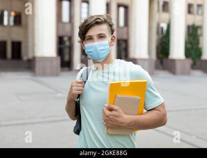 Student guy wearing face mask posing with books, standing outdoors near university building, copy space Stock Photo