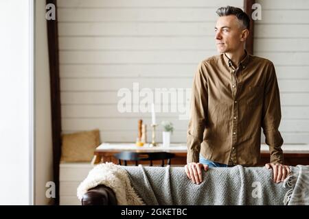 Smiling middle aged white man standing in the modern living room, leaning on a couch, looking aside Stock Photo