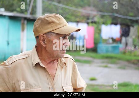 Elderly man standing in rural yard on background of well and hanged laundry. Concept of life in village, old age Stock Photo